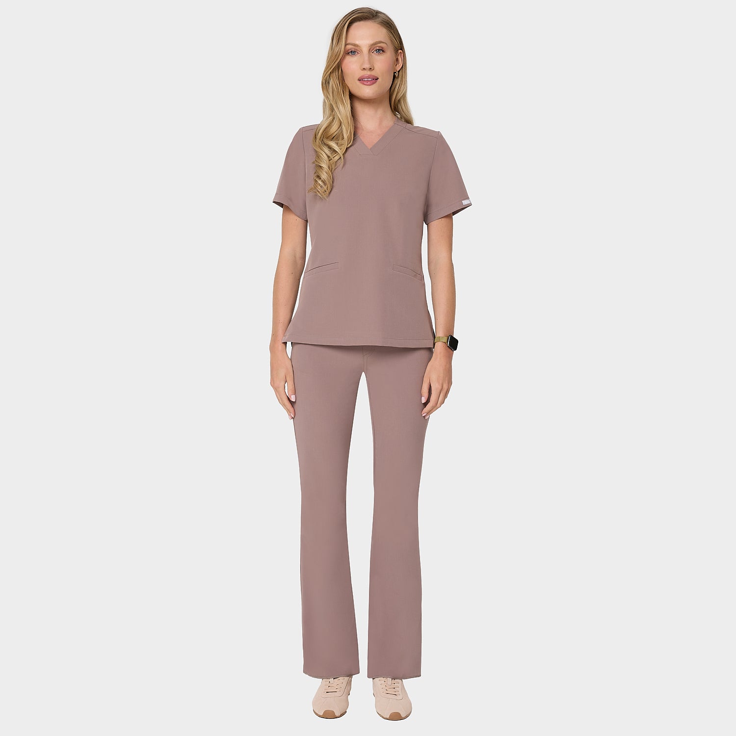 Bluza medyczna Emily w kolorze LIGHT BROWN od Vena Uniformy.
Modelka stoi na jasnym tle w  komplecie medycznym: bluza z krótkim rękawem oraz spodnie flare, zestawione z beżowymi sneakersami.