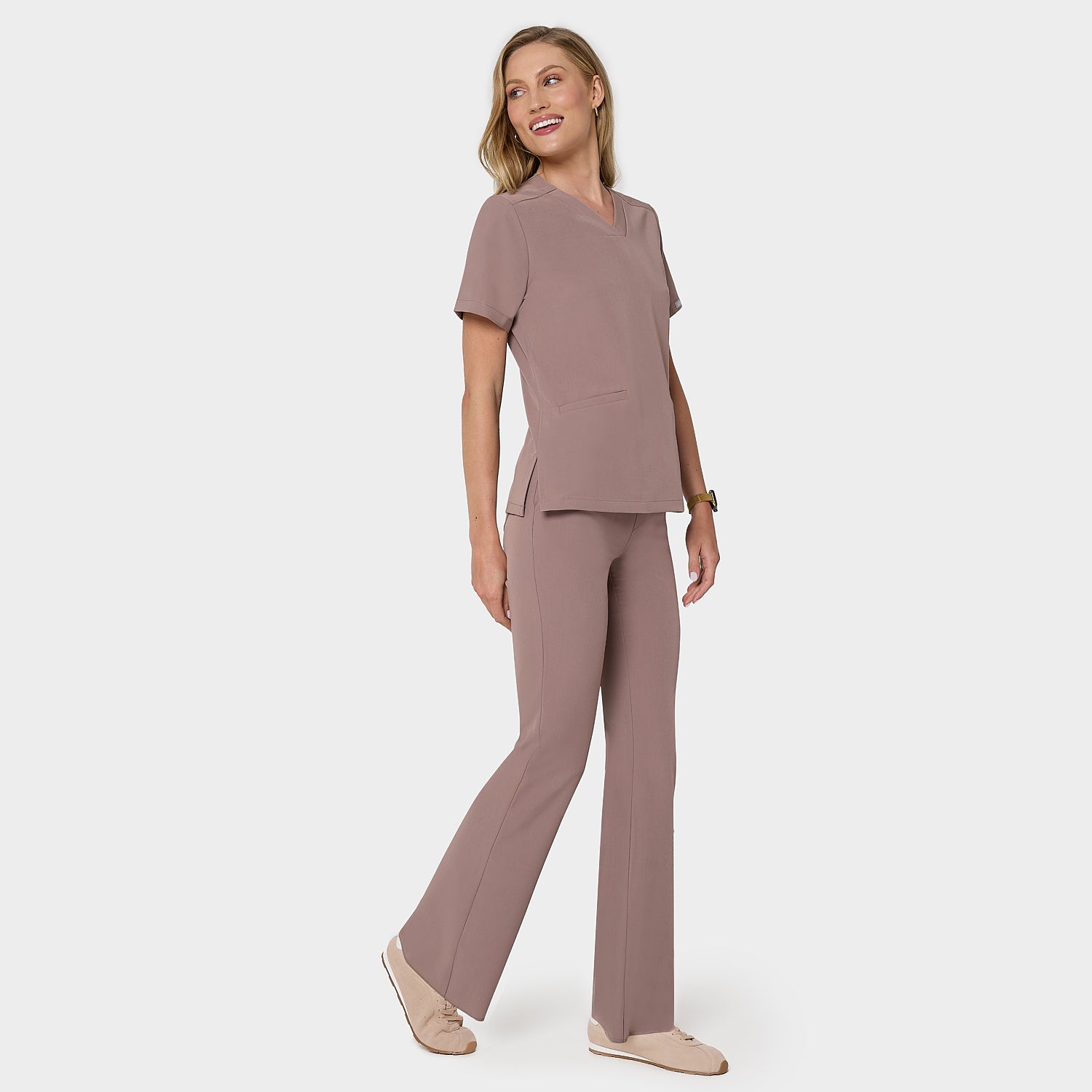 Bluza medyczna Emily w kolorze LIGHT BROWN od Vena Uniformy.
Modelka stoi na jasnym tle w  komplecie medycznym: bluza z krótkim rękawem oraz spodnie flare, zestawione z beżowymi sneakersami.