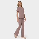 Bluza medyczna Emily w kolorze LIGHT BROWN od Vena Uniformy.
Modelka stoi na jasnym tle w  komplecie medycznym: bluza z krótkim rękawem oraz spodnie flare, zestawione z beżowymi sneakersami.