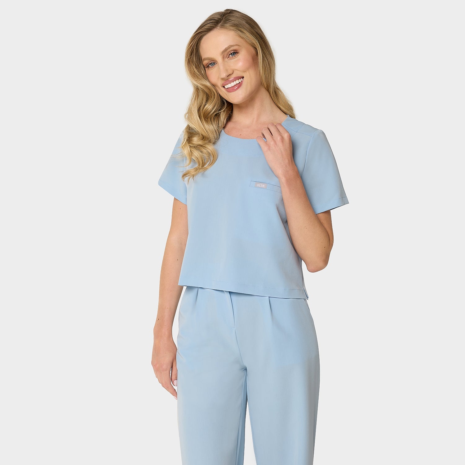 Bluza medyczna Gaja crop w kolorze BABY BLUE od Vena Uniformy.
Modelka stoi na jasnym tle w pastelowym komplecie medycznym: bluza z krótkim rękawem oraz szerokie spodnie.