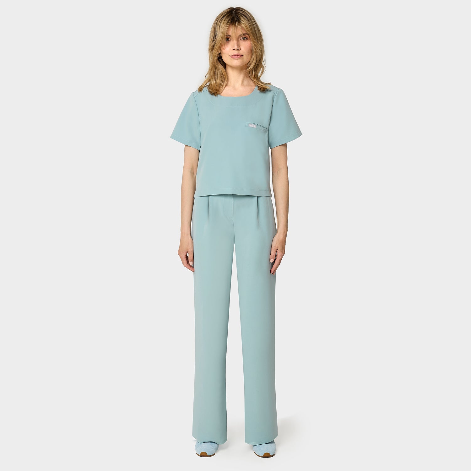 Bluza medyczna Gaja crop w kolorze CLOUDY BLUE od Vena Uniformy.
Modelka stoi na jasnym tle w pastelowym komplecie medycznym: bluza z krótkim rękawem oraz szerokie spodnie, zestawione z niebieskimi sneakersami.
