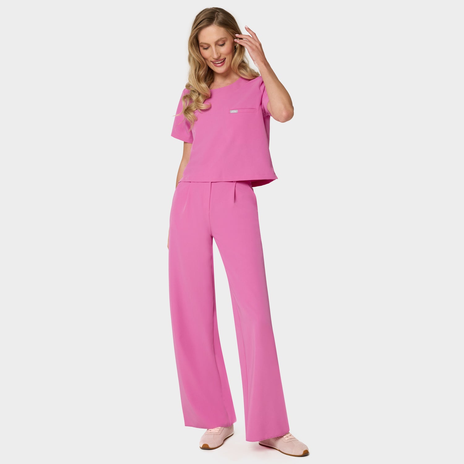 Bluza medyczna Gaja crop w kolorze Disco Pink od Vena Uniformy.
Modelka na jasnym tle w komplecie medycznym: bluza z krótkim rękawem oraz szerokie spodnie, zestawione z beżowymi sneakersami.