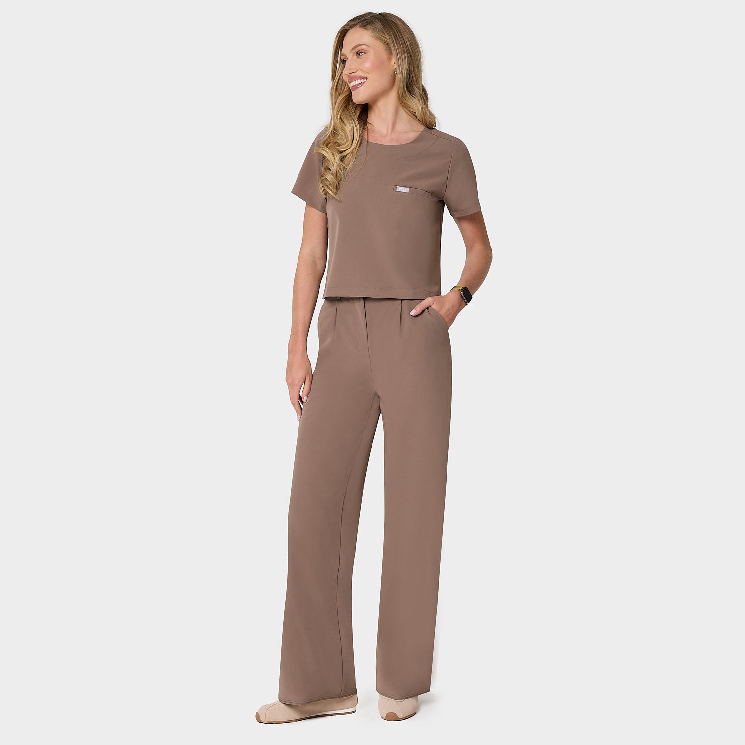 Bluza medyczna Gaja crop w kolorze MOCHA od Vena Uniformy.
Modelka stoi na jasnym tle w  komplecie medycznym: bluza z krótkim rękawem oraz szerokie spodnie, zestawione z beżowymi sneakersami.
