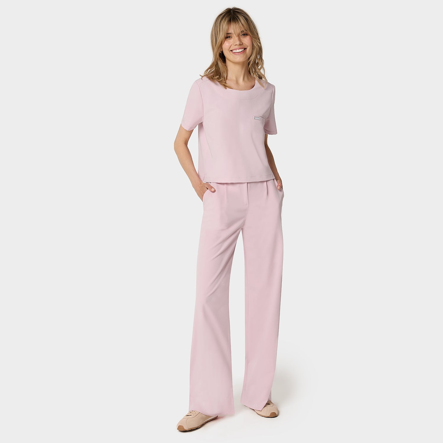 Bluza medyczna Gaja crop w kolorze PASTEL PINK od Vena Uniformy.
Modelka stoi na jasnym tle w pastelowym komplecie medycznym: bluza z krótkim rękawem oraz szerokie spodnie, zestawione z beżowymi sneakersami.
