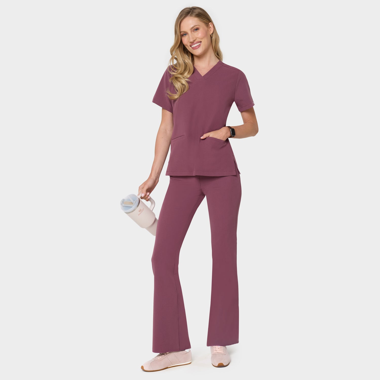 Bluza medyczna Emily w kolorze Dusty Plum od Vena Uniformy.
Modelka stoi na jasnym tle w komplecie medycznym: bluza z krótkim rękawem oraz spodnie flare, zestawione z beżowymi sneakersami.