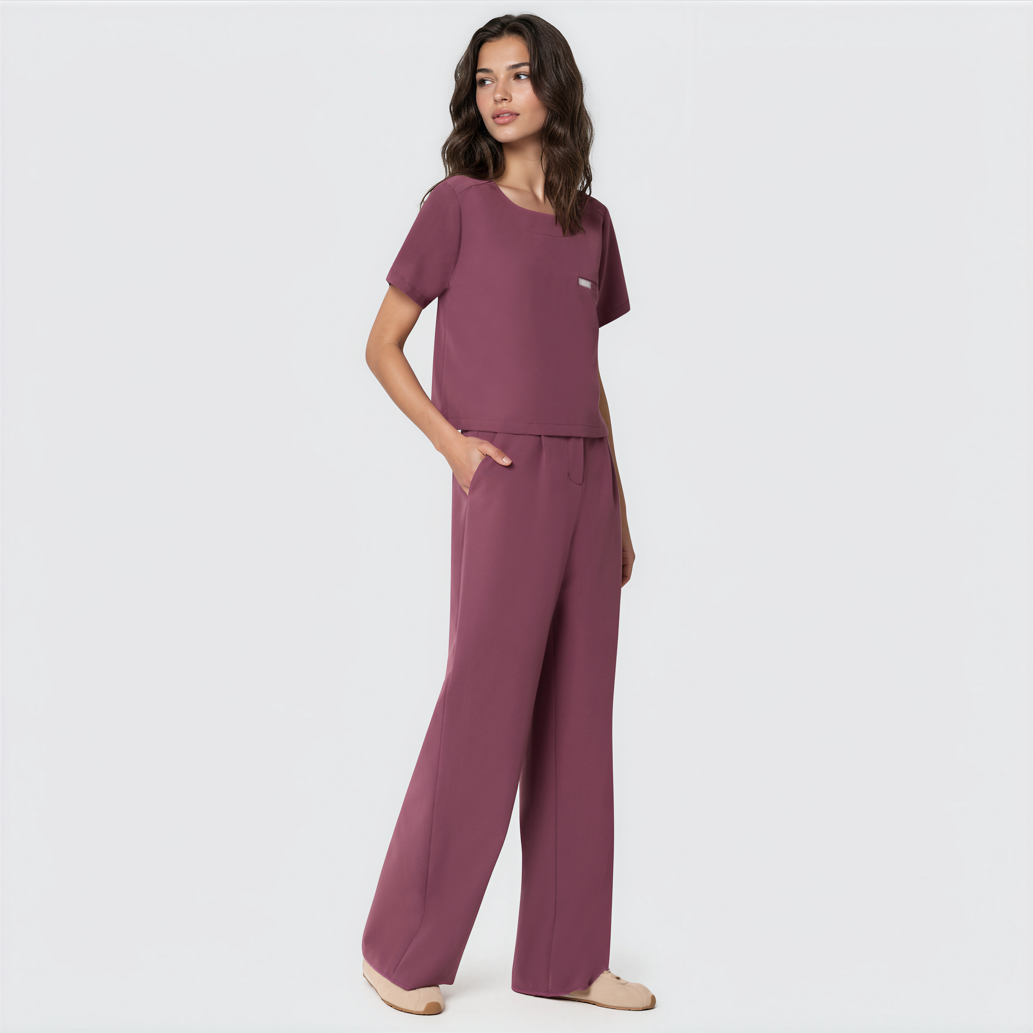 Bluza medyczna Gaja crop w kolorze Dusty Plum od Vena Uniformy.
Modelka na jasnym tle w komplecie medycznym: bluza z krótkim rękawem oraz szerokie spodnie, zestawione z beżowymi sneakersami.